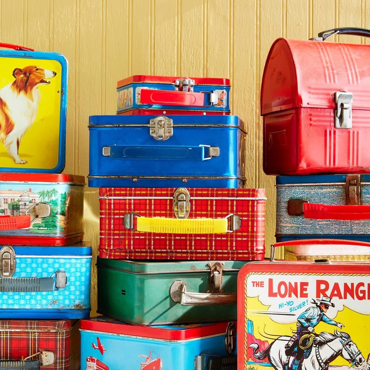 A colorful stack of vintage metal lunchboxes, featuring various designs like a Collie dog, plaid patterns, and “The Lone Ranger,” set against a yellow wooden wall.