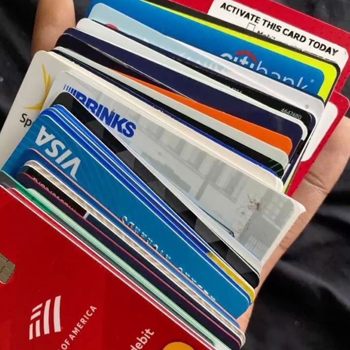 A hand holds a fanned-out stack of various credit and debit cards from different banks, showing multiple colorful card edges and logos, including Visa and Bank of America.