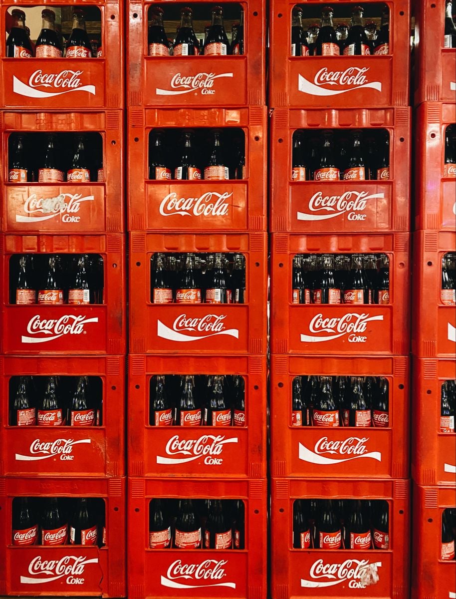 Red plastic crates stacked vertically, each filled with glass Coca-Cola bottles. The crates are branded with white Coca-Cola logos and arranged in a neat, organized pattern, creating a vibrant display.