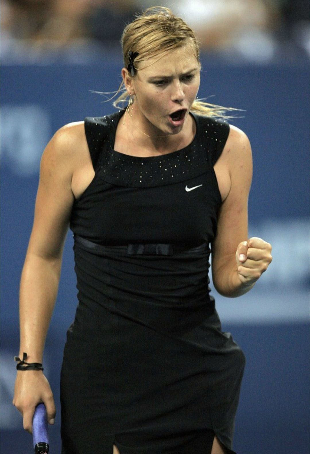 A female tennis player in a black dress clenches her fist and shouts in celebration on the court, holding a racquet in her other hand. The background is blurred, highlighting her intense expression.