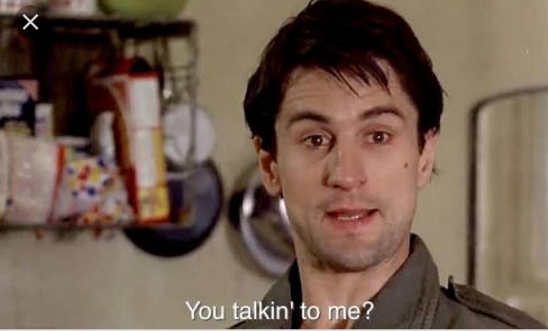A man with short dark hair stands indoors, looking slightly to the side with a serious expression. White text at the bottom reads, "You talkin' to me?" Shelves with various items appear in the background.