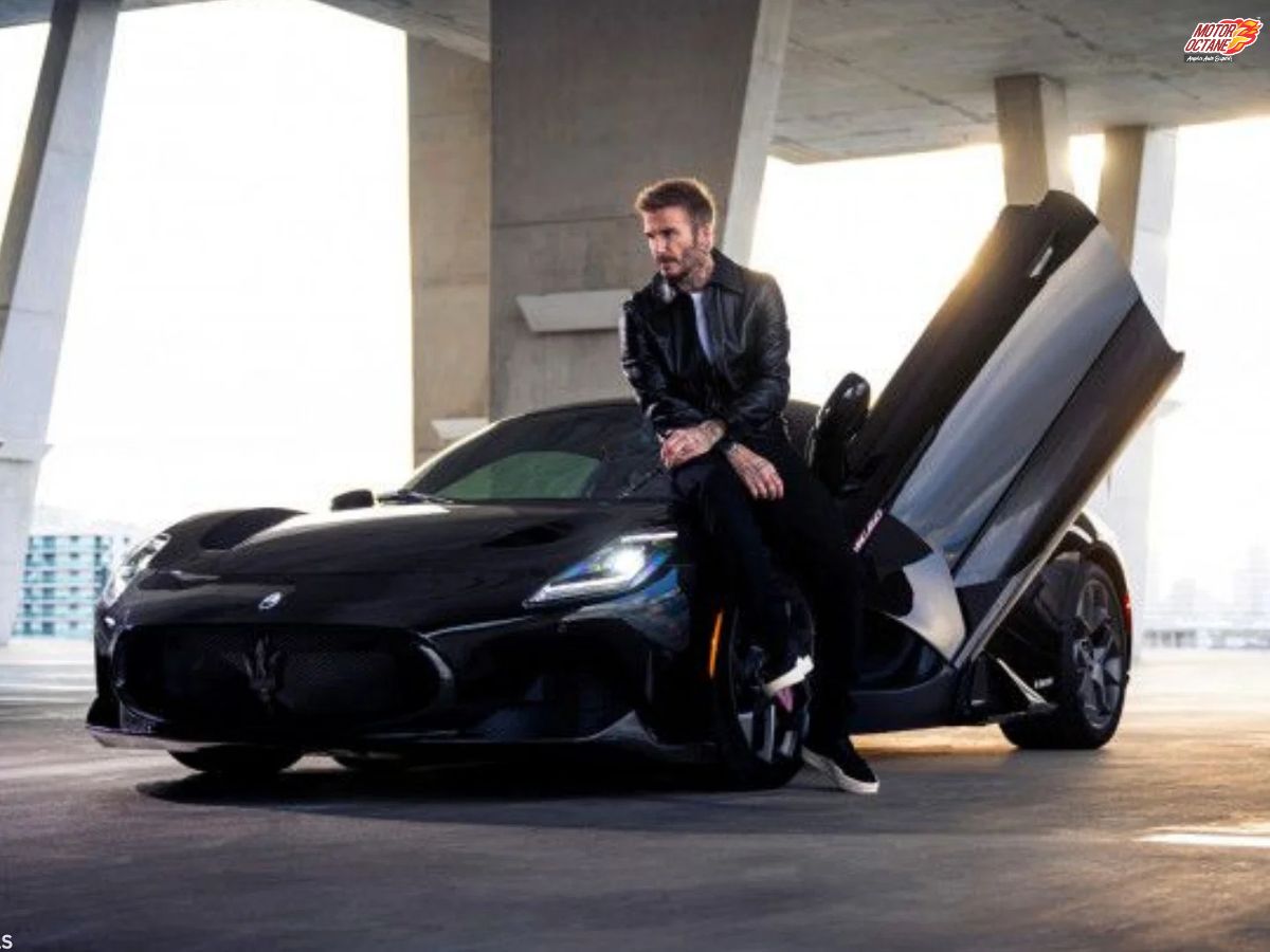 A man in a black jacket sits on the hood of a sleek black sports car with its driver-side door open upward, parked in a modern, sunlit concrete structure.