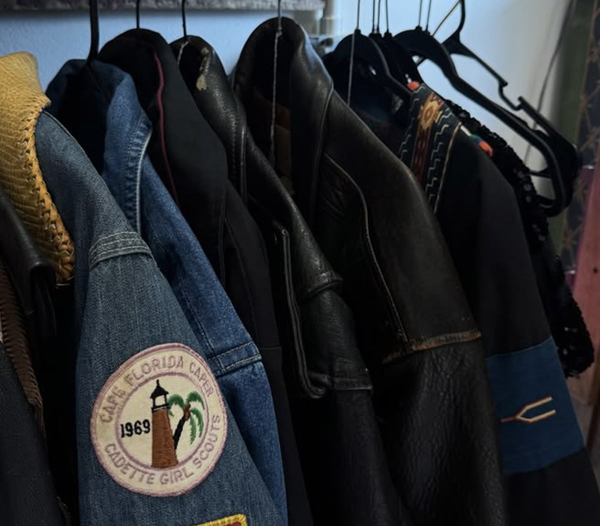 A row of jackets on hangers, including a denim jacket with a round patch that reads "Camp Florida Camp Chibitte Girl Scouts 1969" featuring a palm tree graphic.