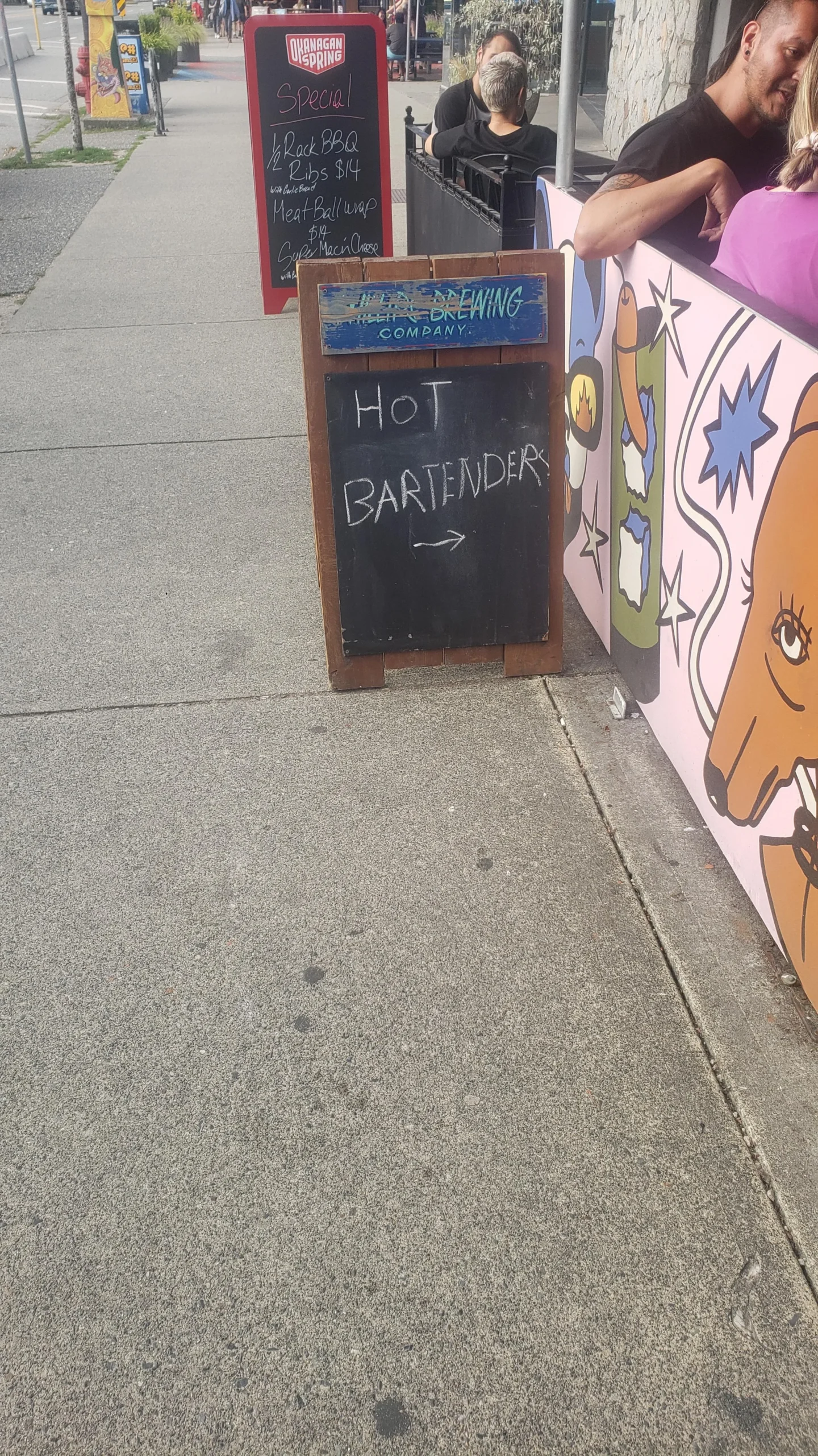 A sidewalk chalkboard sign reads "HOT BARTENDER →" with an arrow pointing right. Behind it, another menu board and outdoor seating area with people and colorful, cartoon-style artwork are visible.