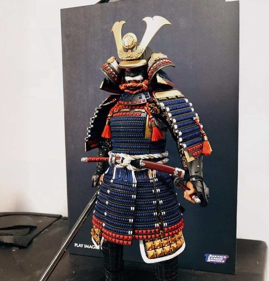 A detailed samurai action figure in blue, red, and gold armor stands against a dark background, holding a sword with both hands. The figure wears a traditional helmet with a gold crest and intricate details.