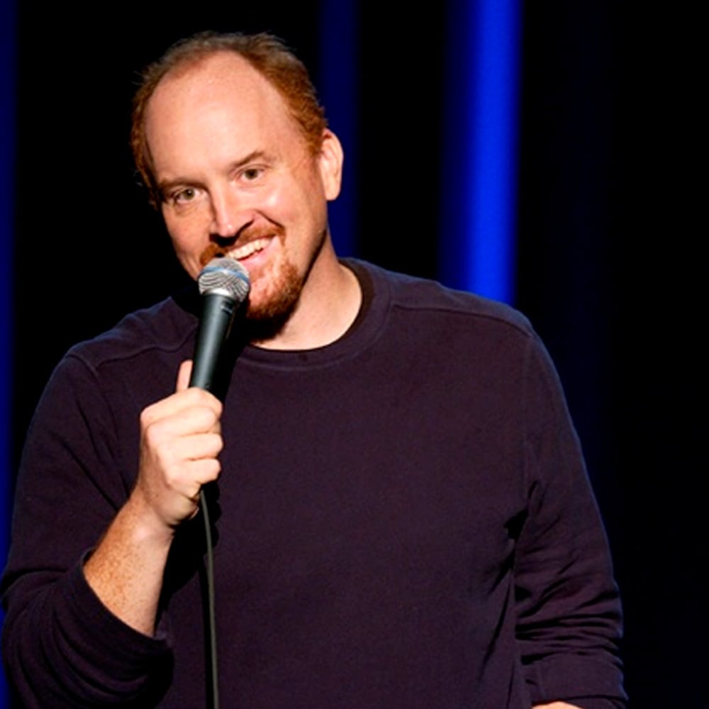 A man with short red hair and a beard smiles while holding a microphone on stage, standing in front of a dark background with blue vertical lights.