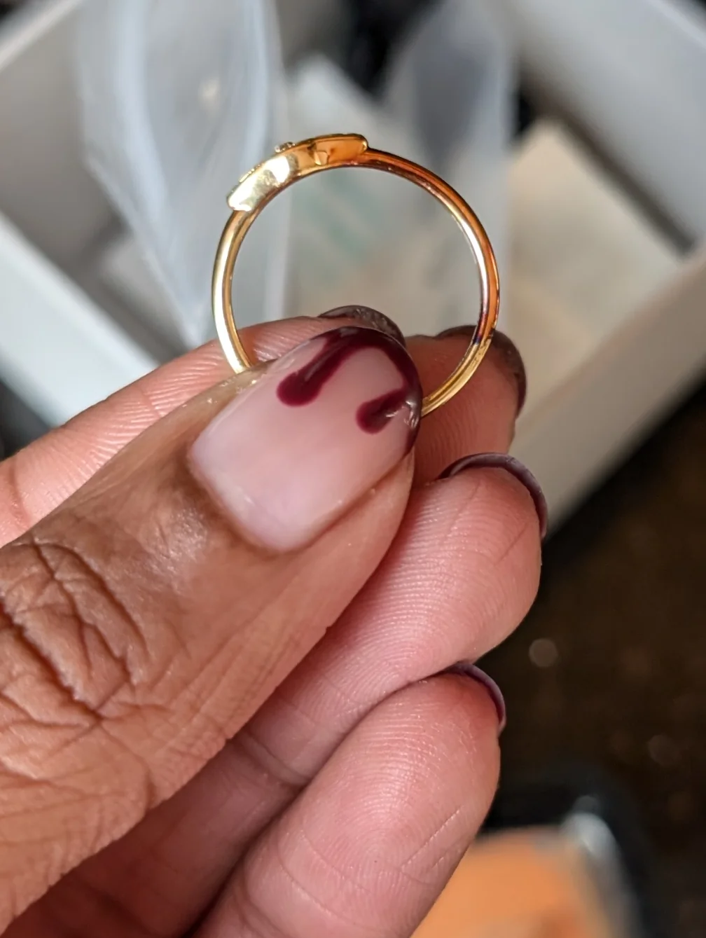 A hand with manicured nails holding a gold ring with a small gap at the top, in front of a blurred background with a white box.