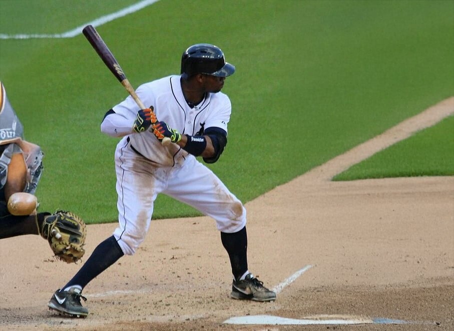 A baseball player in a batting stance at home plate, wearing a white uniform and helmet, prepares to swing as the catcher crouches behind him on a baseball field.