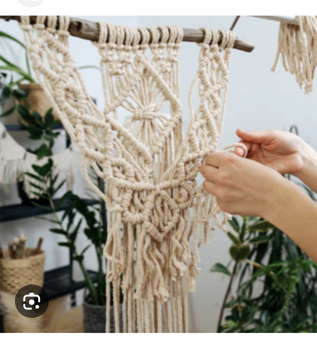 Hands working on a detailed macramé wall hanging made of cream-colored cord, attached to a wooden stick, with green plants and shelves in the background.