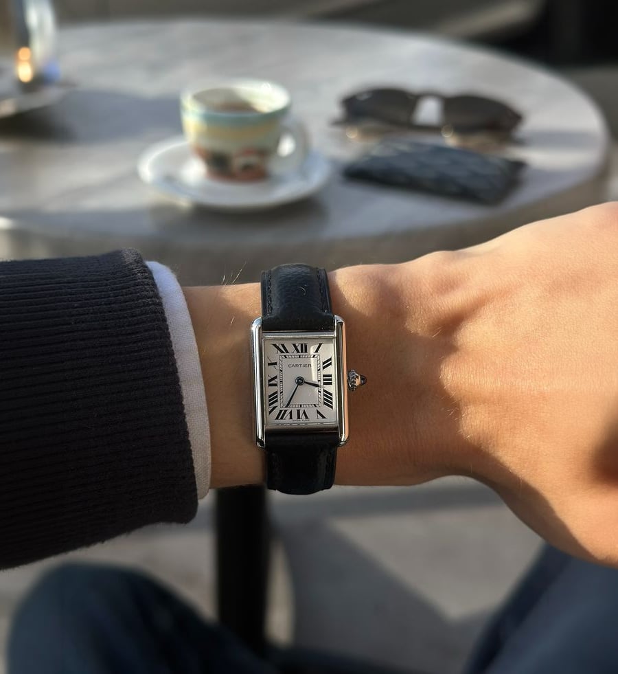 A person wearing a rectangular watch with a black leather strap and Roman numerals on the dial, sitting at a table with a cup, saucer, sunglasses, and wallet in the background.