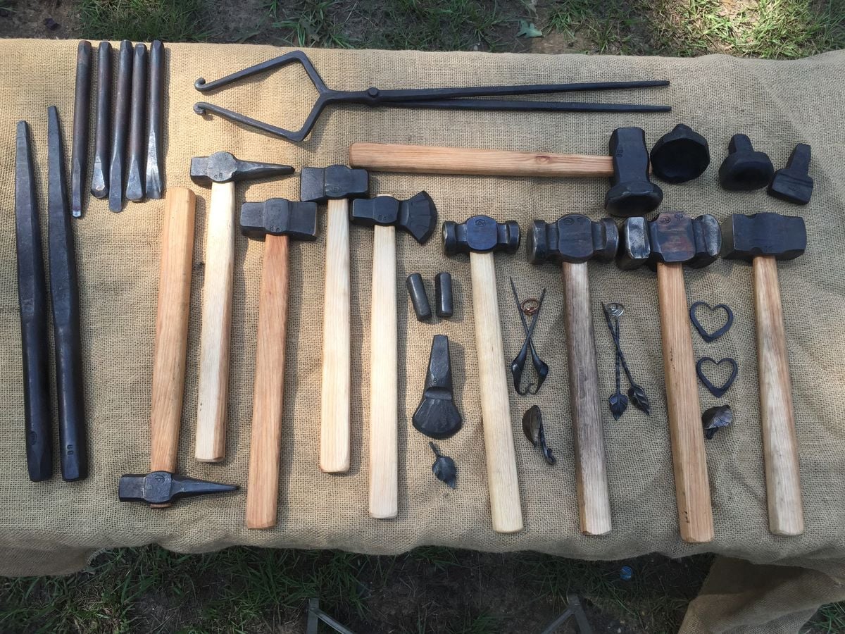 A variety of blacksmith tools, including hammers, tongs, chisels, punches, and finished metal pieces, laid out neatly on a burlap-covered table outdoors.