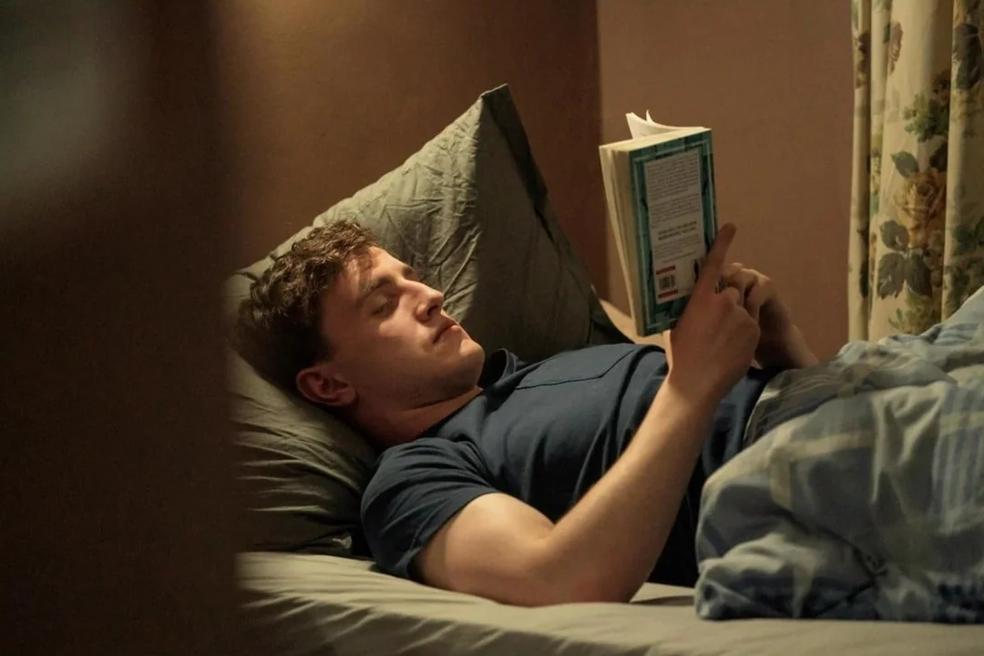 A person lies on a bed under a gray blanket, propped up on pillows, reading a book. The room has soft lighting and floral curtains are visible in the background.