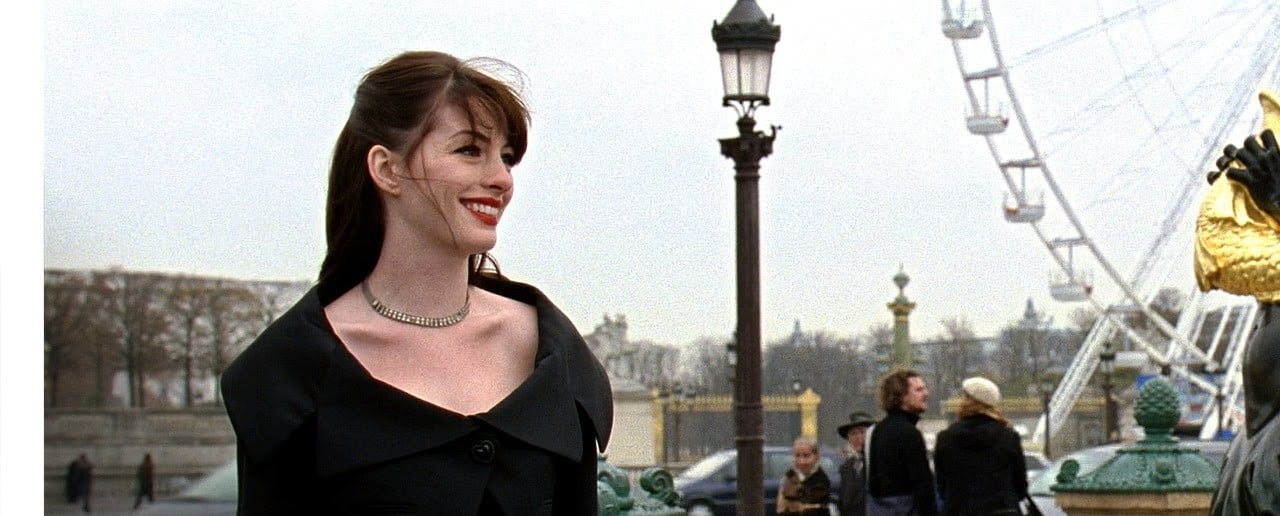 A woman with brown hair and red lipstick, wearing a black dress and a diamond necklace, smiles while standing outdoors. In the background, people walk near a Ferris wheel and ornate lamp posts.