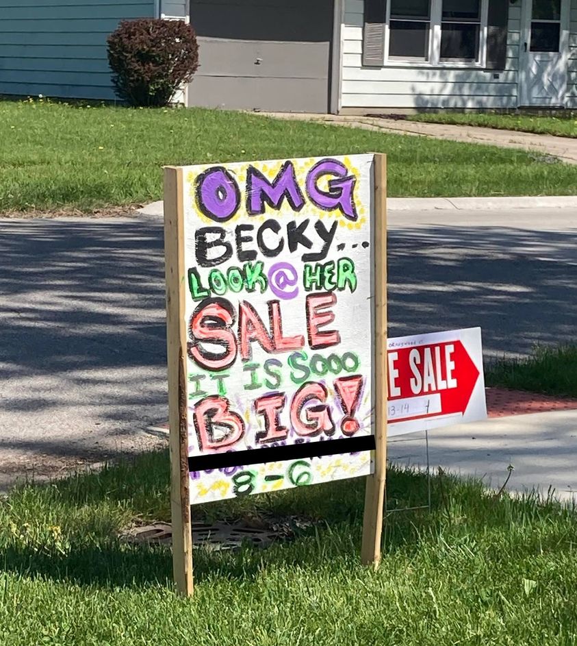 A colorful yard sign reads, “OMG BECKY… LOOK @ HER SALE IT IS SOOO BIG!” with decorative lettering, placed on a lawn near a street. Another red and white "For Sale" sign is visible in the background.