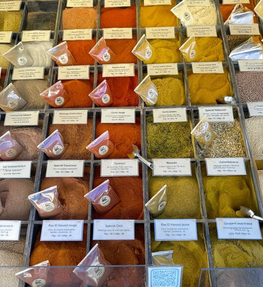 A display of assorted spices in clear containers and labeled plastic bags at a market, showing a colorful variety of powders and blends, including curry, chili, and masala.