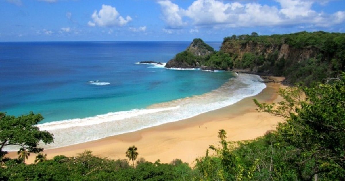 A scenic view of a tropical beach with golden sand, turquoise water, gentle waves, and lush green cliffs under a partly cloudy blue sky.