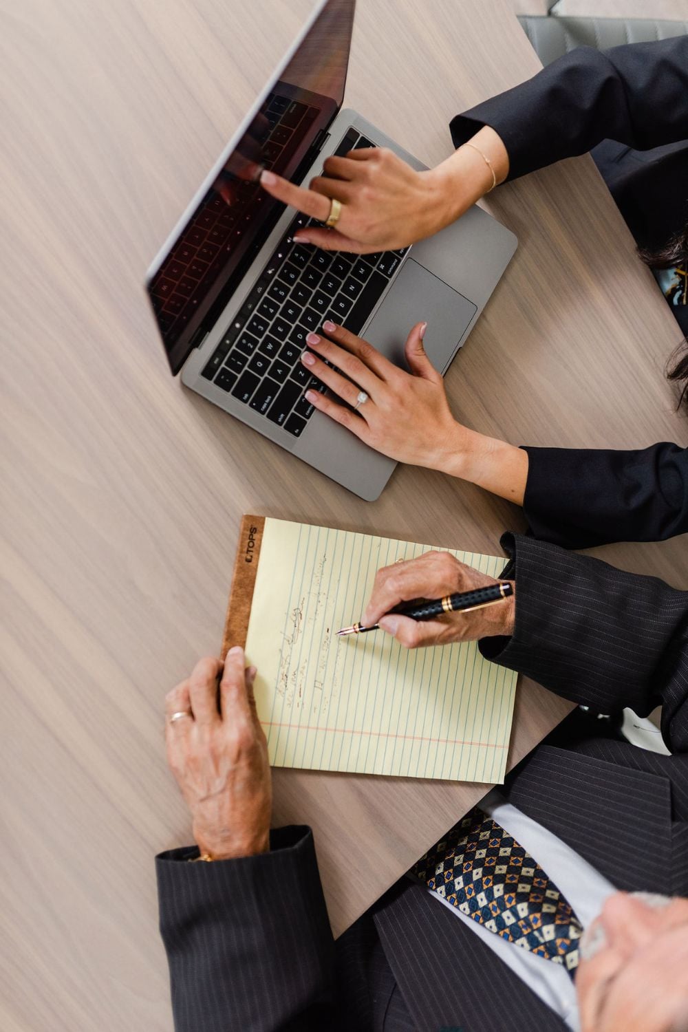 Two people at a desk, one typing on a laptop and pointing at the screen, the other writing notes on a yellow legal pad. Both are wearing business attire.