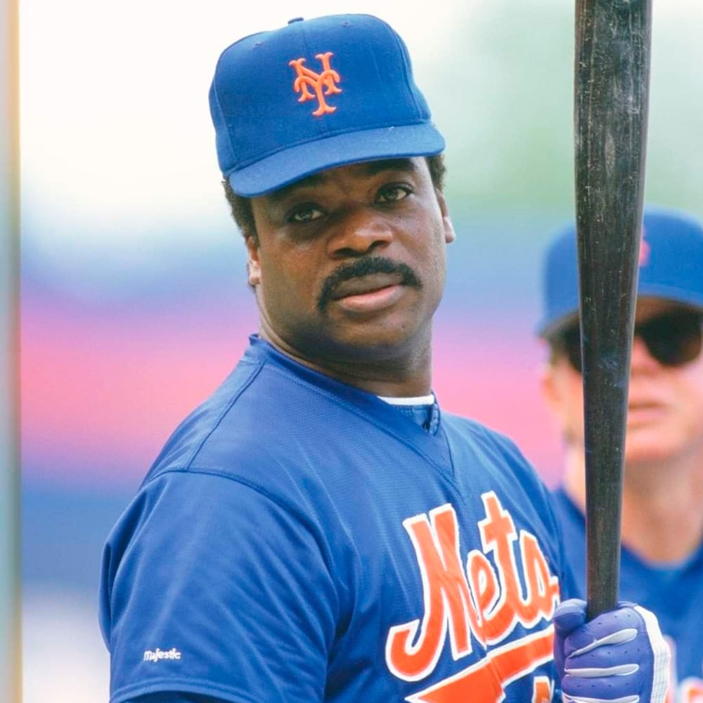 A baseball player in a blue New York Mets uniform and cap holds a bat, looking toward the camera. Another person in a cap and sunglasses is visible in the background.