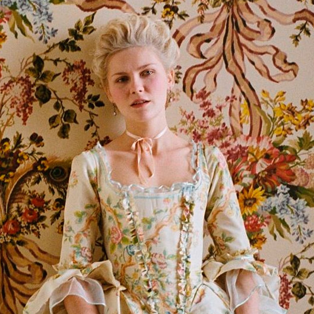 A woman with pale blond hair styled in an 18th-century updo wears a pastel floral Rococo dress and a pink ribbon choker, posing against a colorful, ornate floral wallpaper.