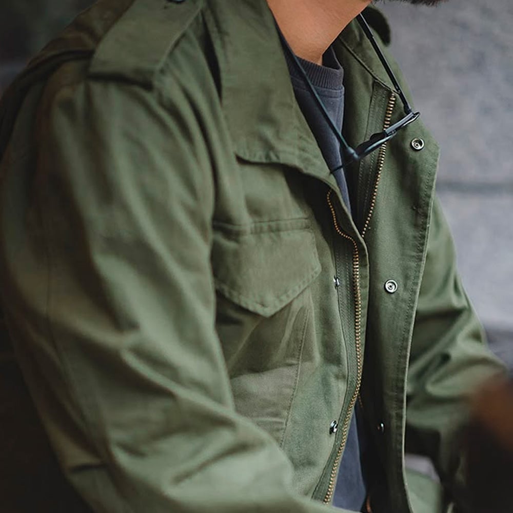A person wearing a green military-style jacket over a grey shirt, with sunglasses hanging from the jacket’s collar. Only the lower face and upper body are visible.