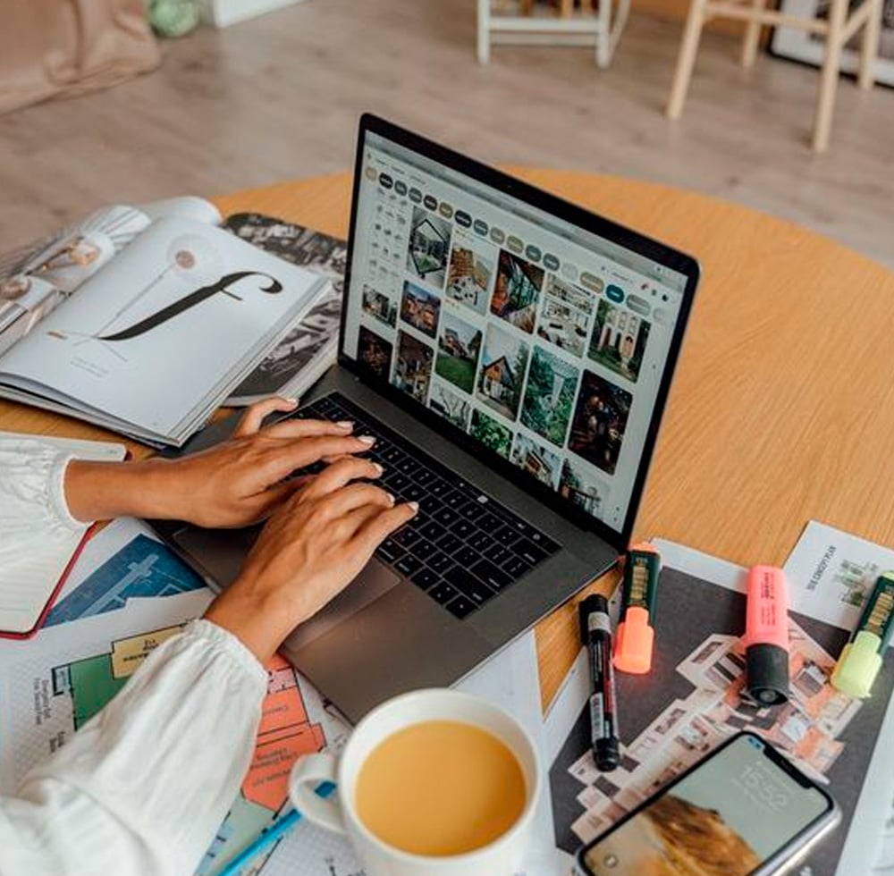 A person types on a laptop displaying a photo gallery. The desk holds magazines, highlighters, markers, papers, a smartphone, and a mug of coffee or tea. The background shows a wooden floor and chairs.