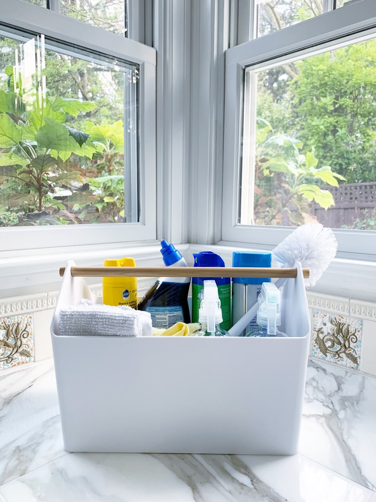 A white cleaning caddy filled with supplies like spray bottles, sponges, cloths, and a scrub brush sits on a marble countertop in a bright corner by large windows with a garden view outside.