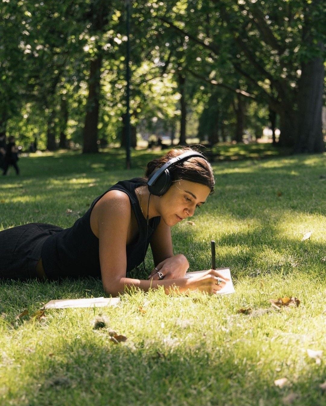 A person wearing headphones lies on their stomach on the grass in a sunlit park, writing or drawing in a notebook. Trees and dappled sunlight fill the background, creating a peaceful, relaxed atmosphere.