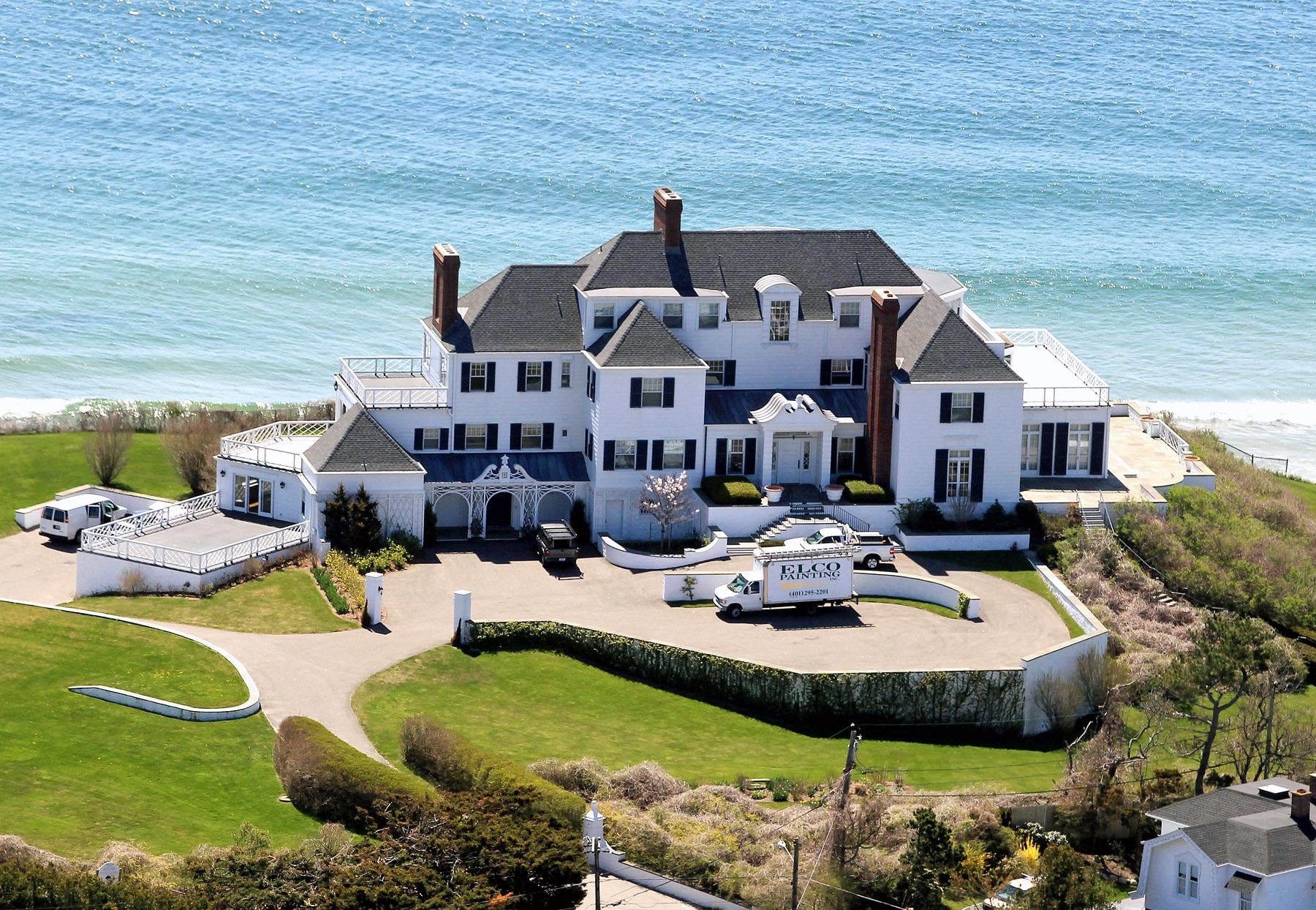 A large white mansion with black roofs sits on a green lawn by the ocean. A helicopter labeled "RELAX" is parked in the driveway near the entrance, and the house overlooks the blue water with waves near the shore.