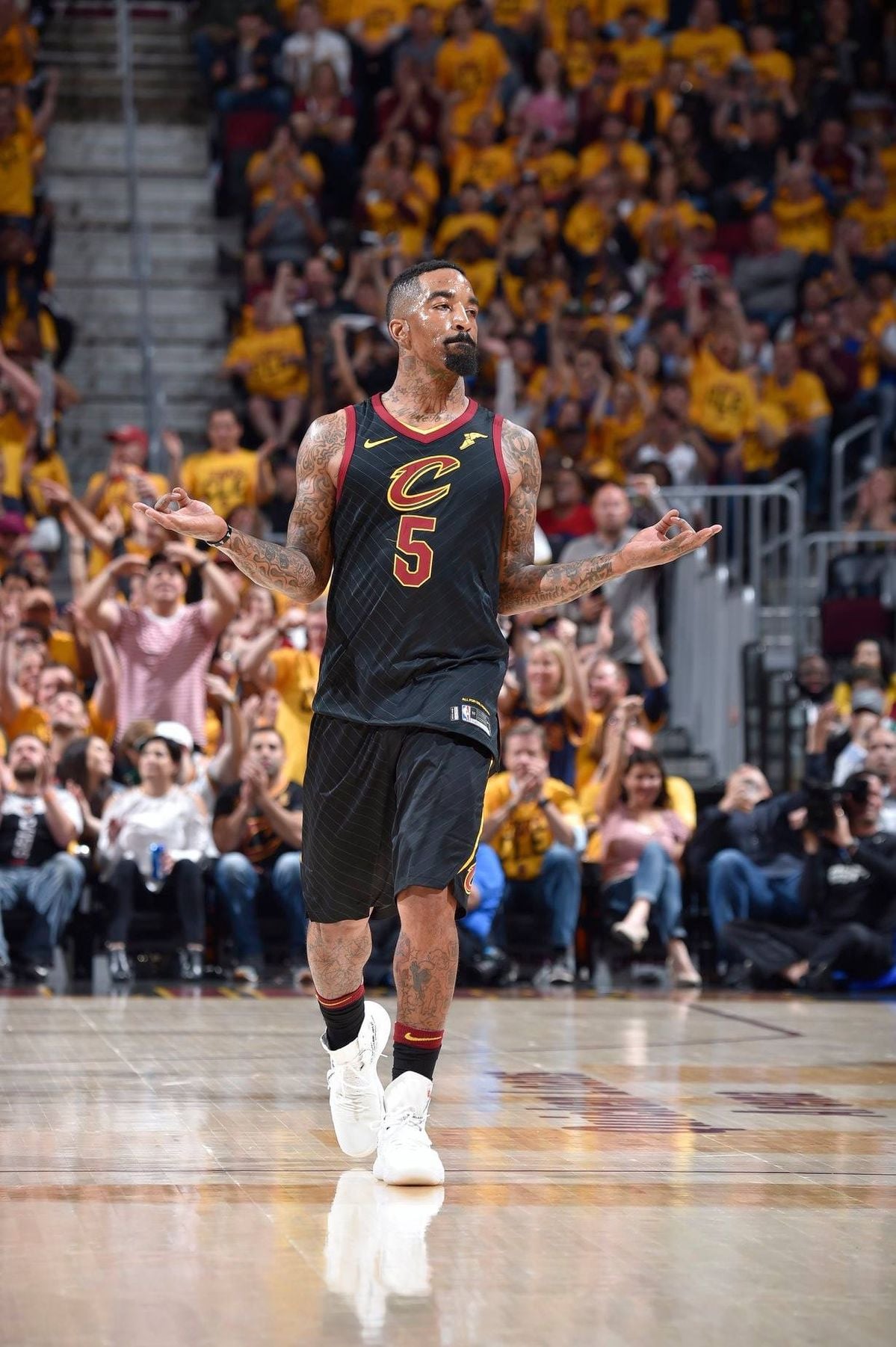 A basketball player in a black Cleveland Cavaliers uniform (#5) stands on the court with arms outstretched, palms up, as fans in yellow cheer in the background during a game.