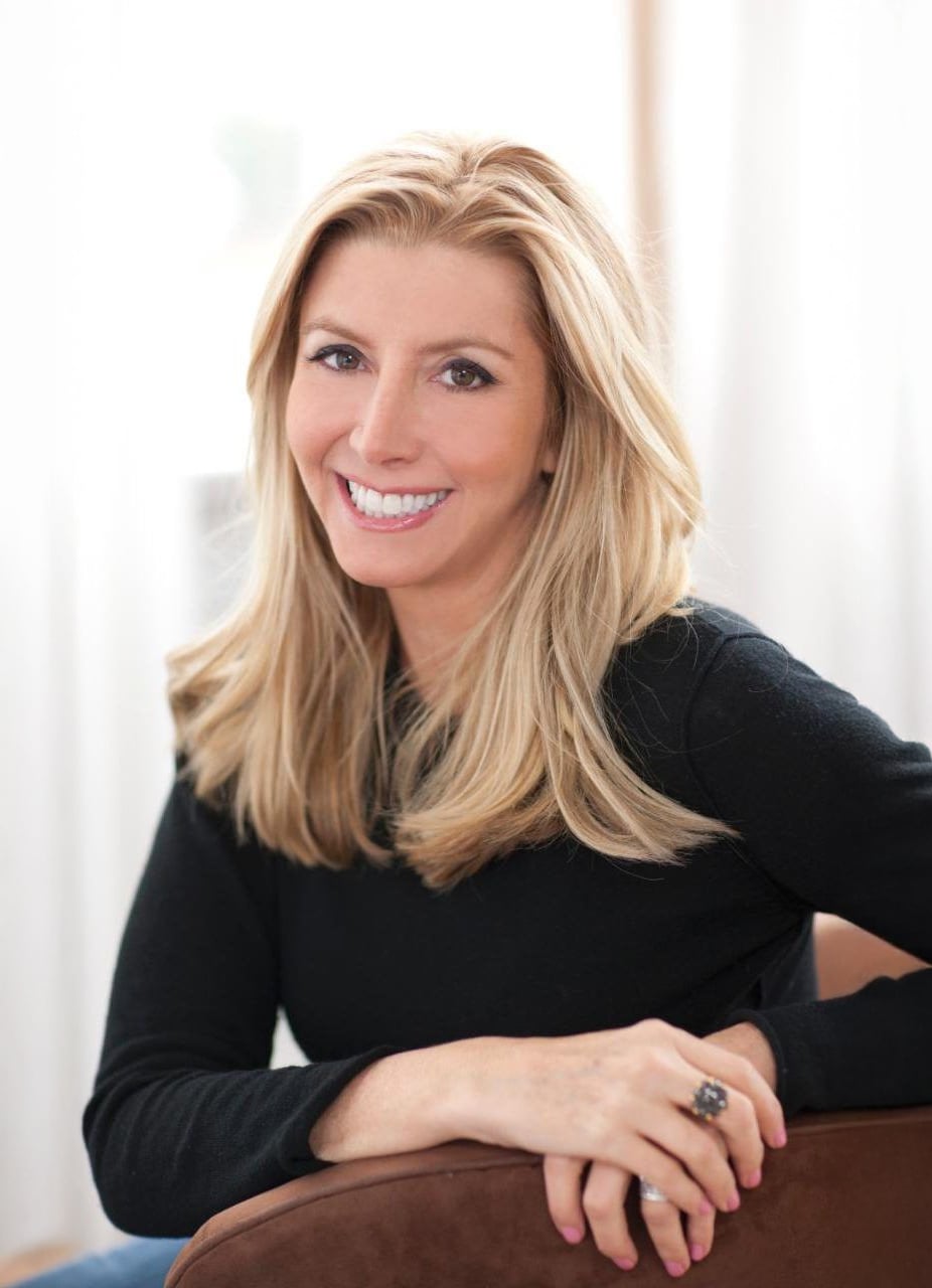 A woman with long blonde hair, wearing a black top, sits on a brown chair and smiles at the camera. She has light pink nail polish and a large ring on her finger. Bright, softly lit background.
