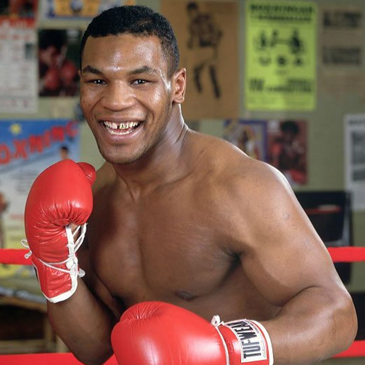 A smiling male boxer stands in a boxing ring, wearing red gloves and no shirt. Colorful posters hang on the wall behind him.