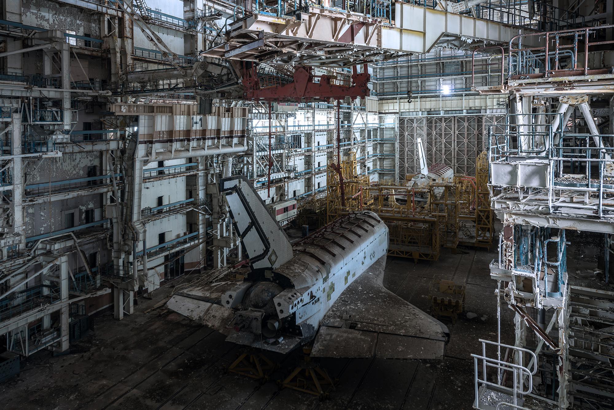 Two abandoned Soviet space shuttles sit inside a large, dilapidated hangar filled with rusted scaffolding, metal beams, and scattered debris under dim natural light.