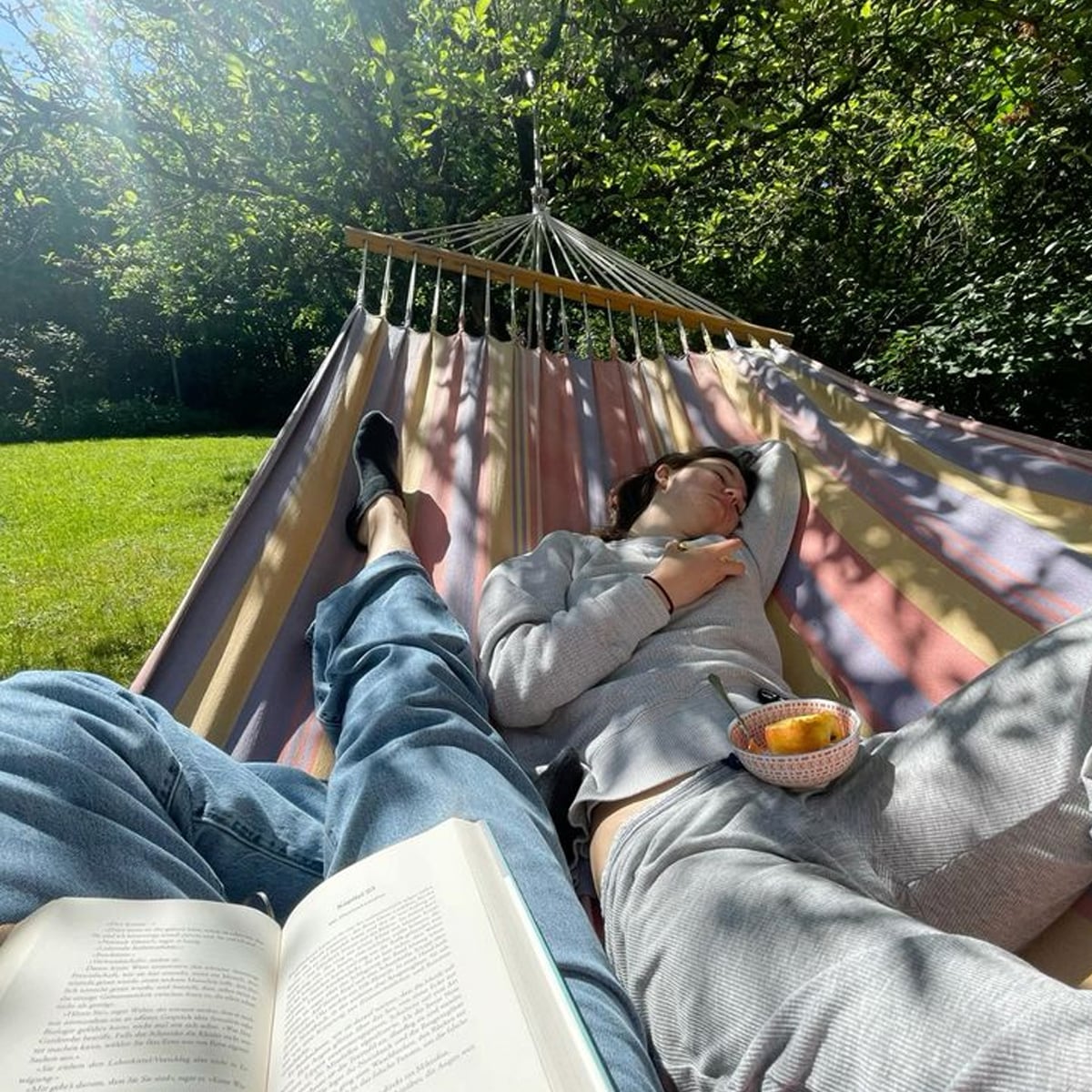 Two people relax on a striped hammock in a sunlit garden. One person is reading a book while the other naps, holding a small bowl of snacks. Green trees and grass surround the hammock, creating a peaceful scene.