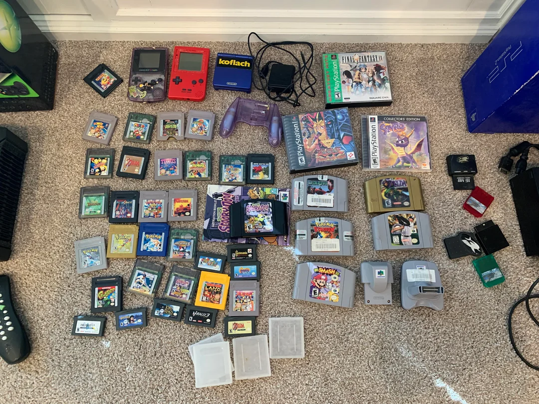 A collection of retro video games and consoles, including Game Boy, Game Boy Advance, Nintendo 64 cartridges, a red Game Boy Color, controllers, chargers, and various game cases arranged on a beige carpet.