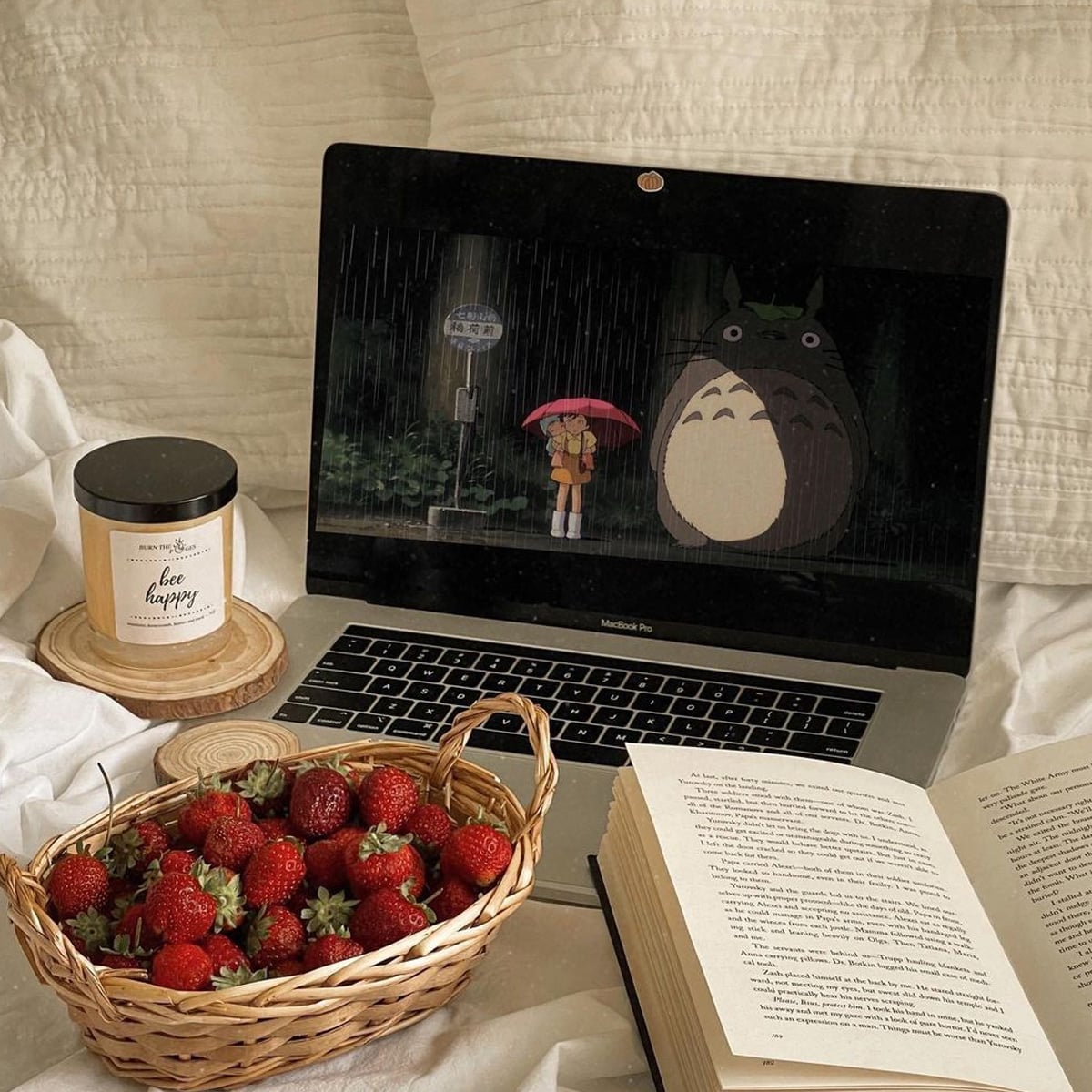 A cozy scene with a laptop showing a Totoro animation, a basket of strawberries, a lit candle, and an open book, all arranged on a bed with white bedding.