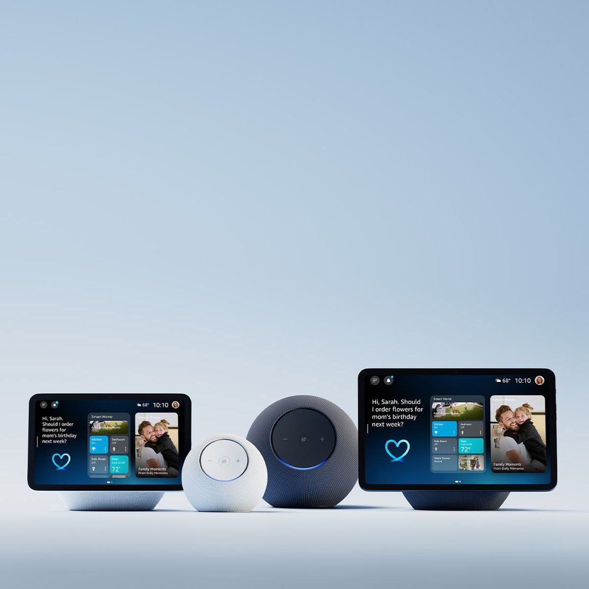 Four smart home devices are displayed on a light surface: two touchscreen smart displays showing home screens with photos and two spherical smart speakers, one small and white, one larger and dark gray, against a blue background.