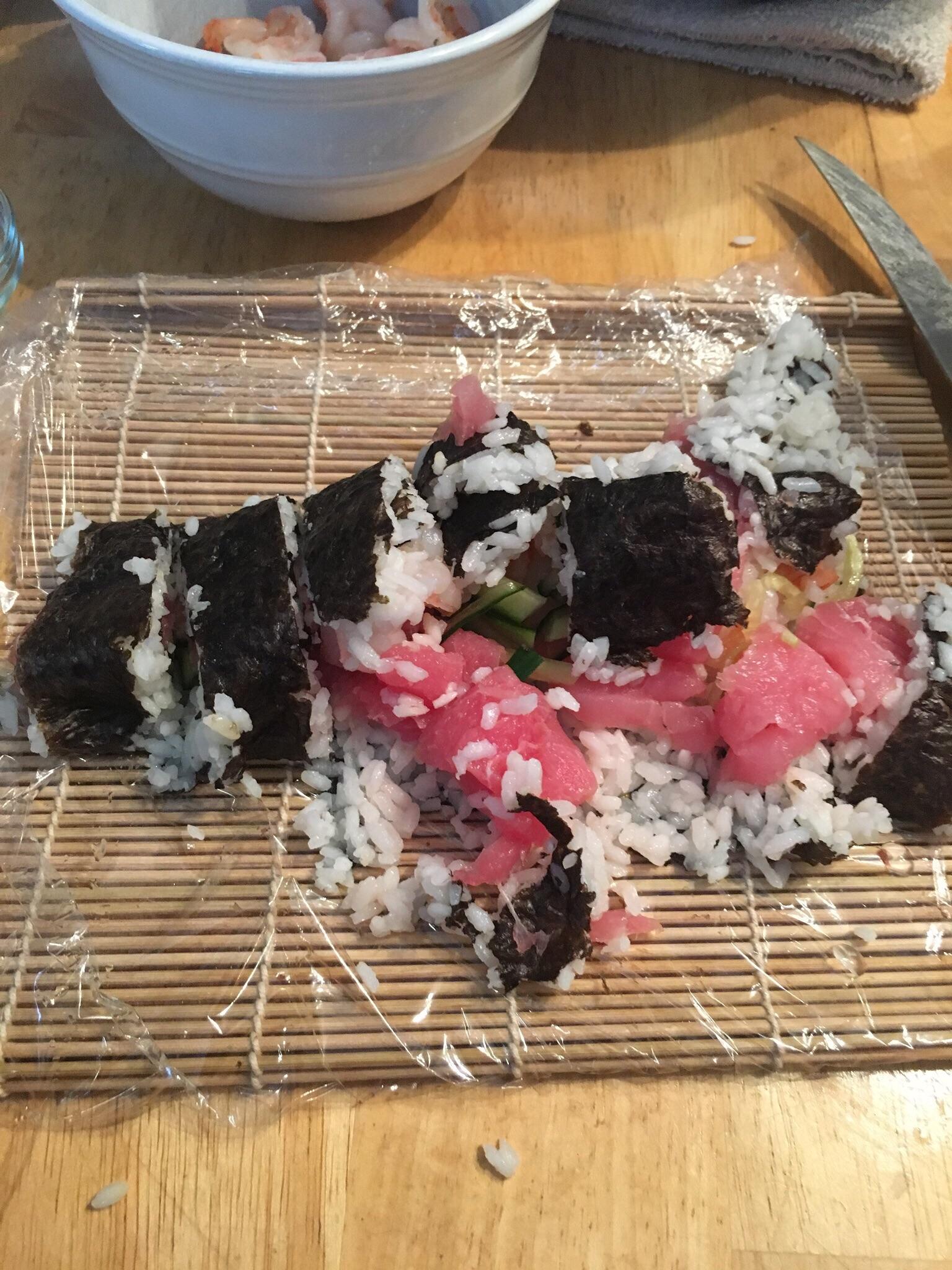 A partially unrolled sushi roll with rice, seaweed, and pink fish sits on a bamboo sushi mat covered in plastic wrap. The sushi pieces are uneven and falling apart. A bowl with shrimp is in the background.