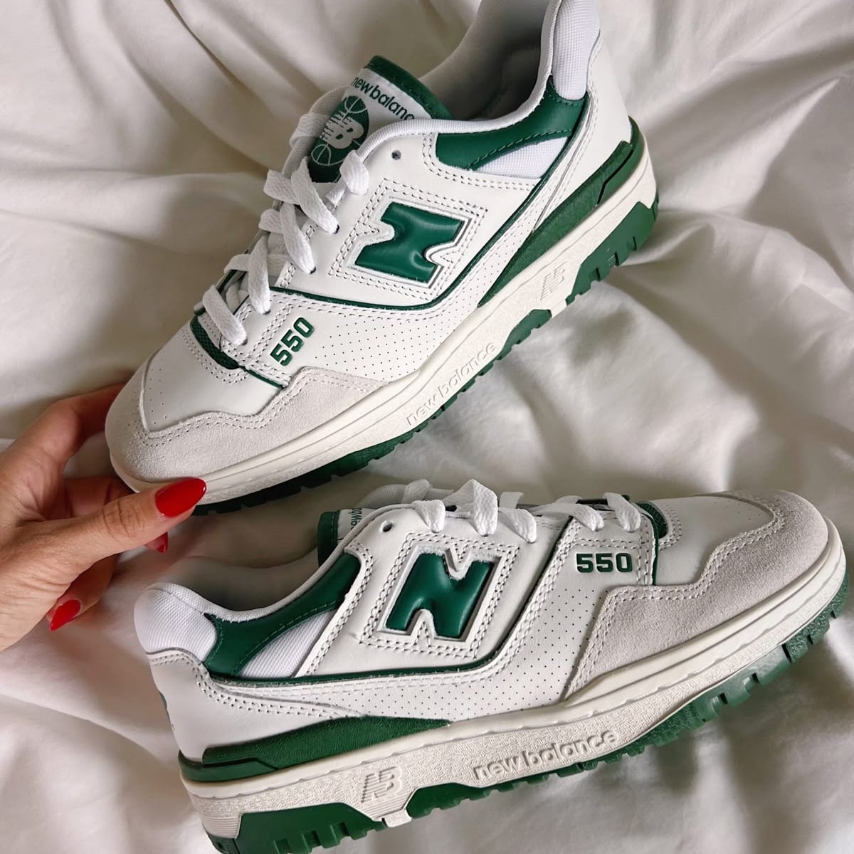 A pair of white and green New Balance 550 sneakers is displayed on a white fabric surface. One shoe is being held, showing the side and top details, including the "N" logo and "550" on the side.
