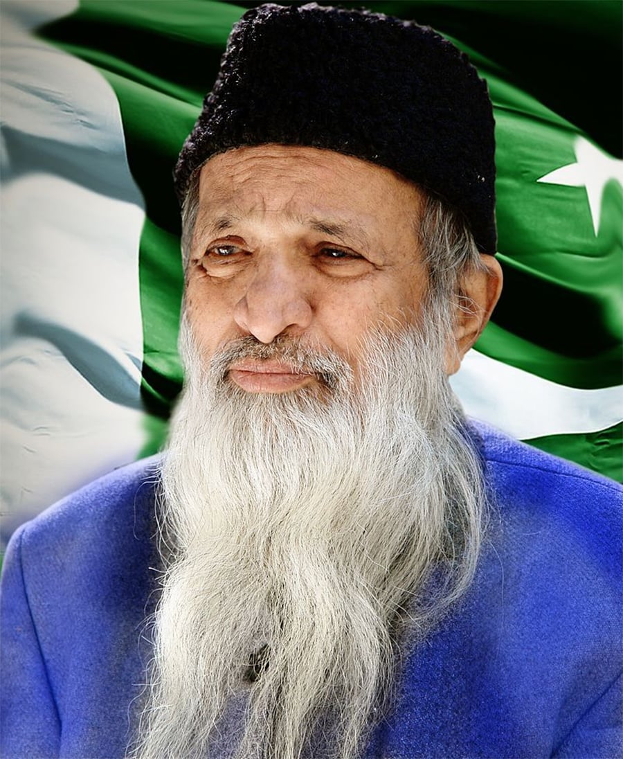 An elderly man with a long white beard, wearing a black cap and blue clothing, stands in front of a waving Pakistan flag.