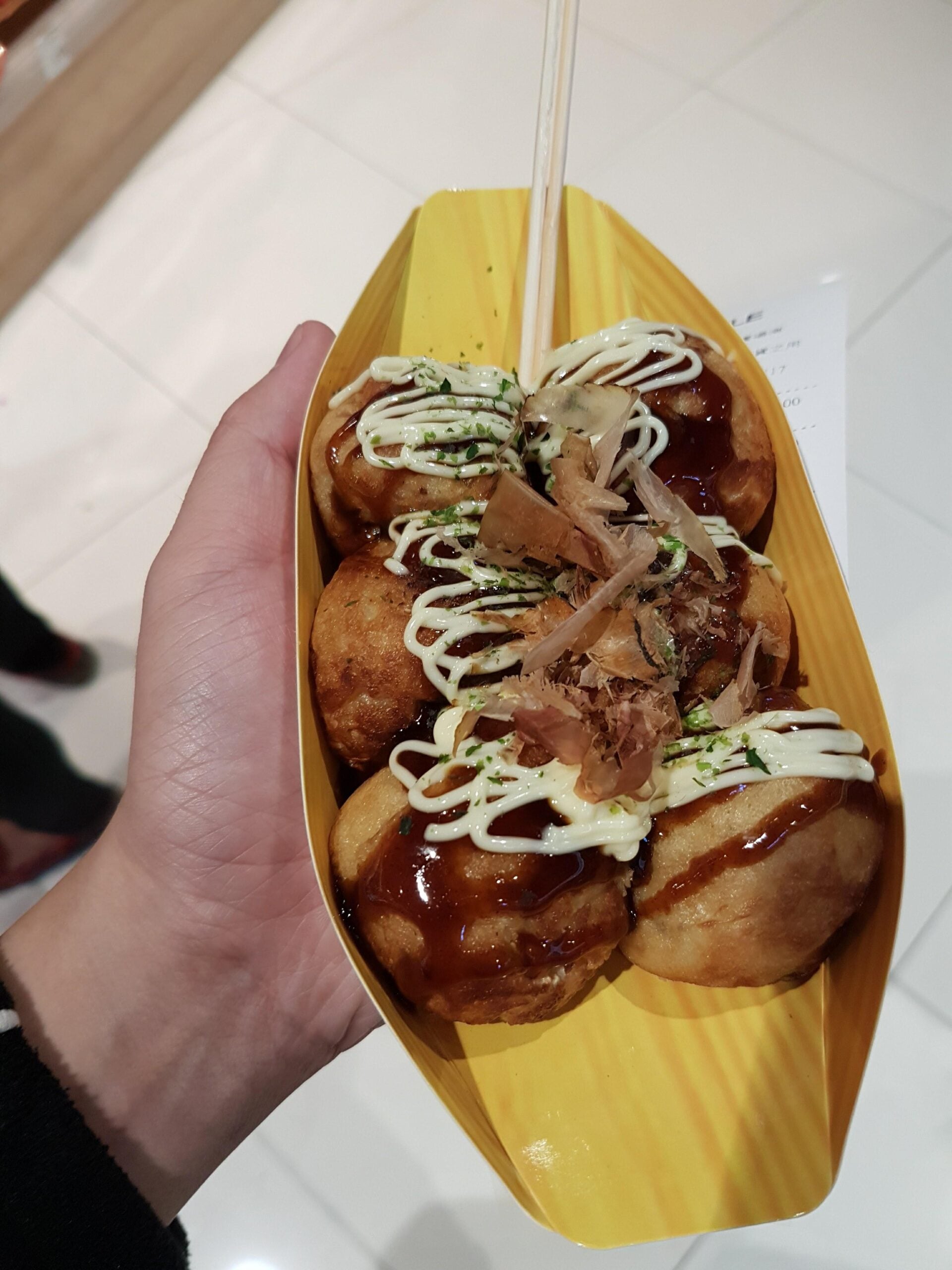 A hand holds a yellow tray with six takoyaki balls topped with mayonnaise, takoyaki sauce, bonito flakes, and green seasoning. Two wooden skewers are inserted for eating. The background is a tiled floor.