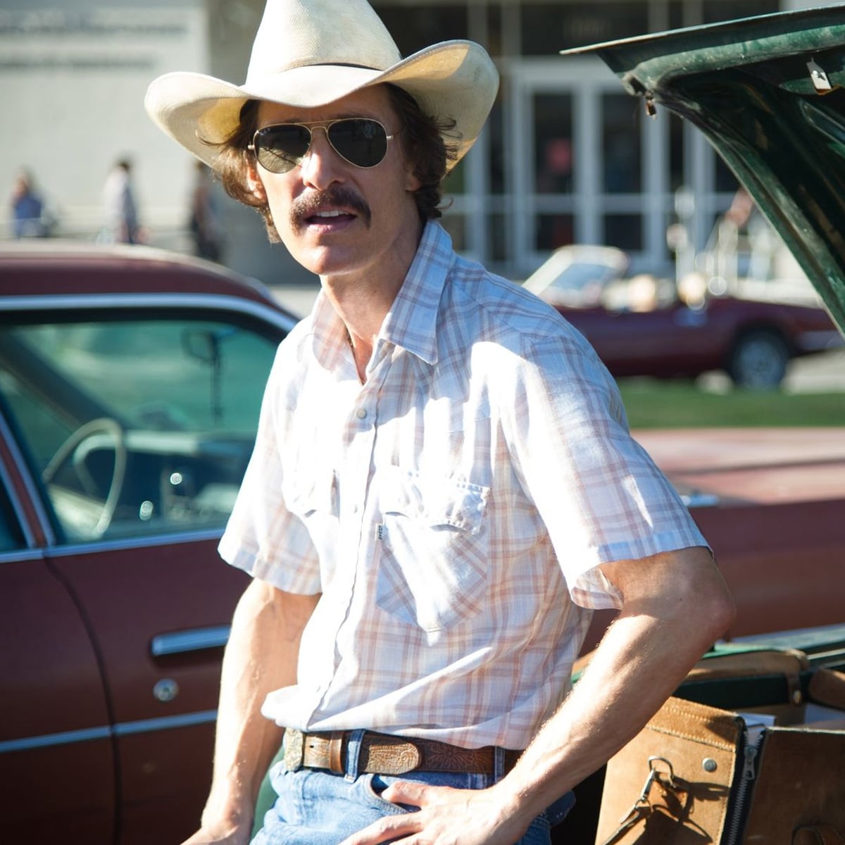 A man in a cowboy hat, aviator sunglasses, and plaid shirt stands beside an open car trunk, with classic cars and a building in the background.