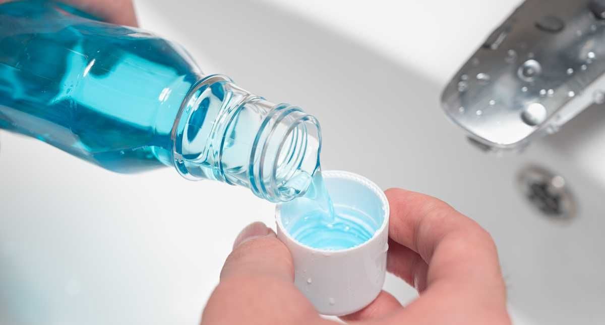 A person pours blue mouthwash from a bottle into a white cap, over a sink with a silver faucet in the background.