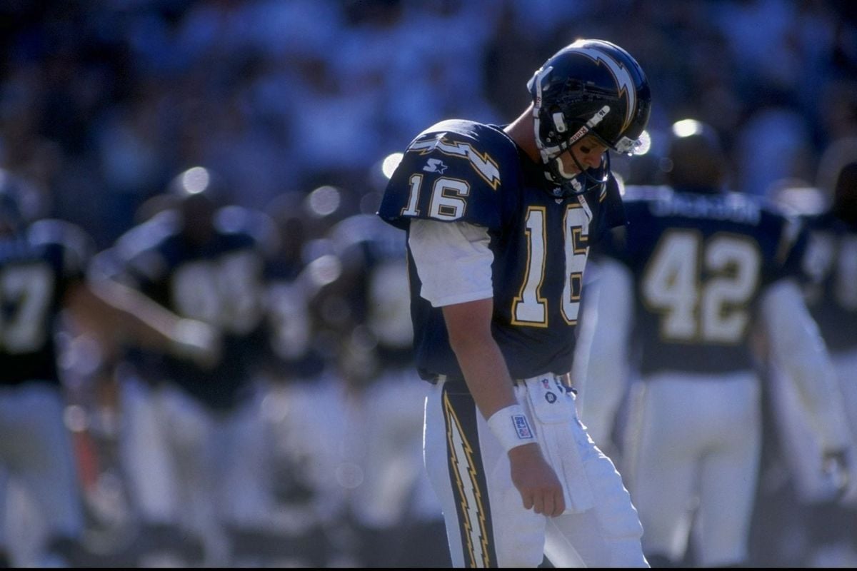 A football player wearing a dark blue San Diego Chargers uniform and helmet, number 16, stands on the field looking down, with teammates and blurred figures in the background.