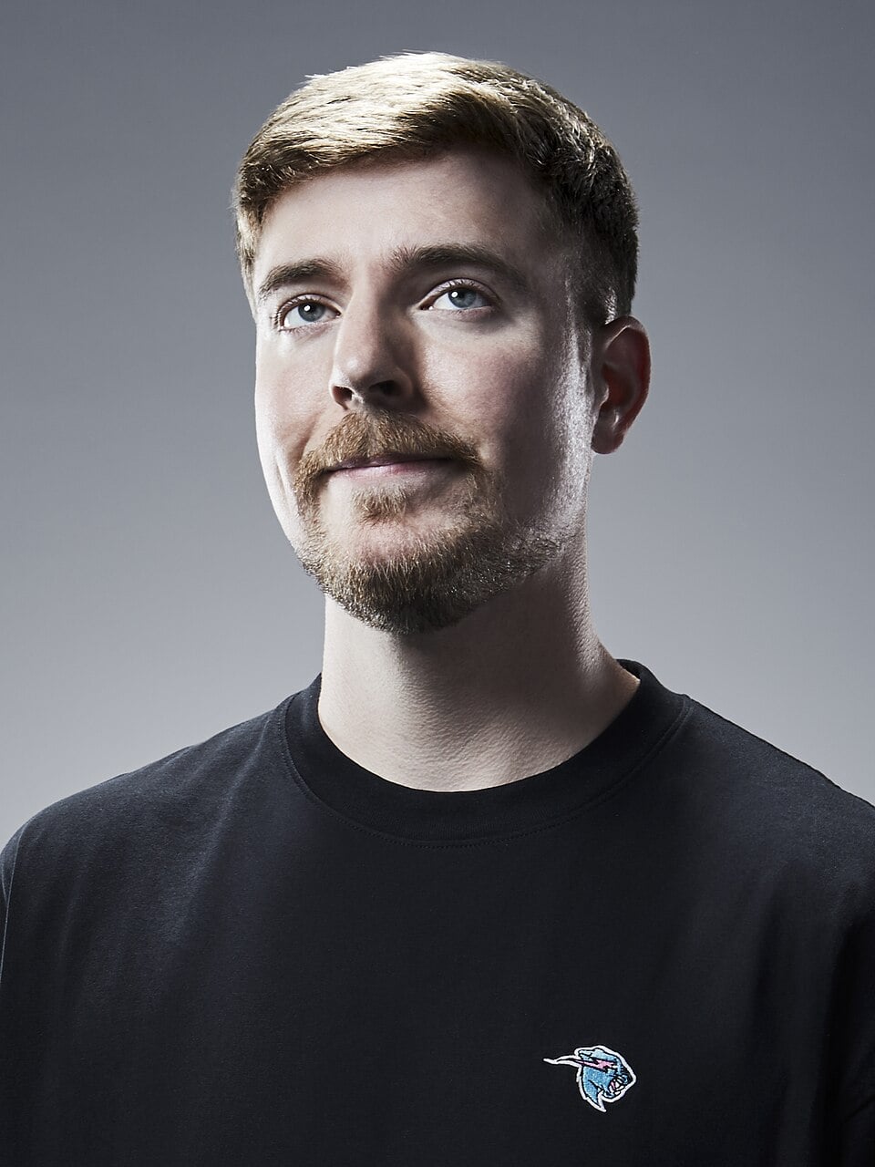 A young man with light brown hair and a short beard, wearing a black t-shirt with a small graphic logo, stands against a neutral gray background, looking upward with a slight smile.