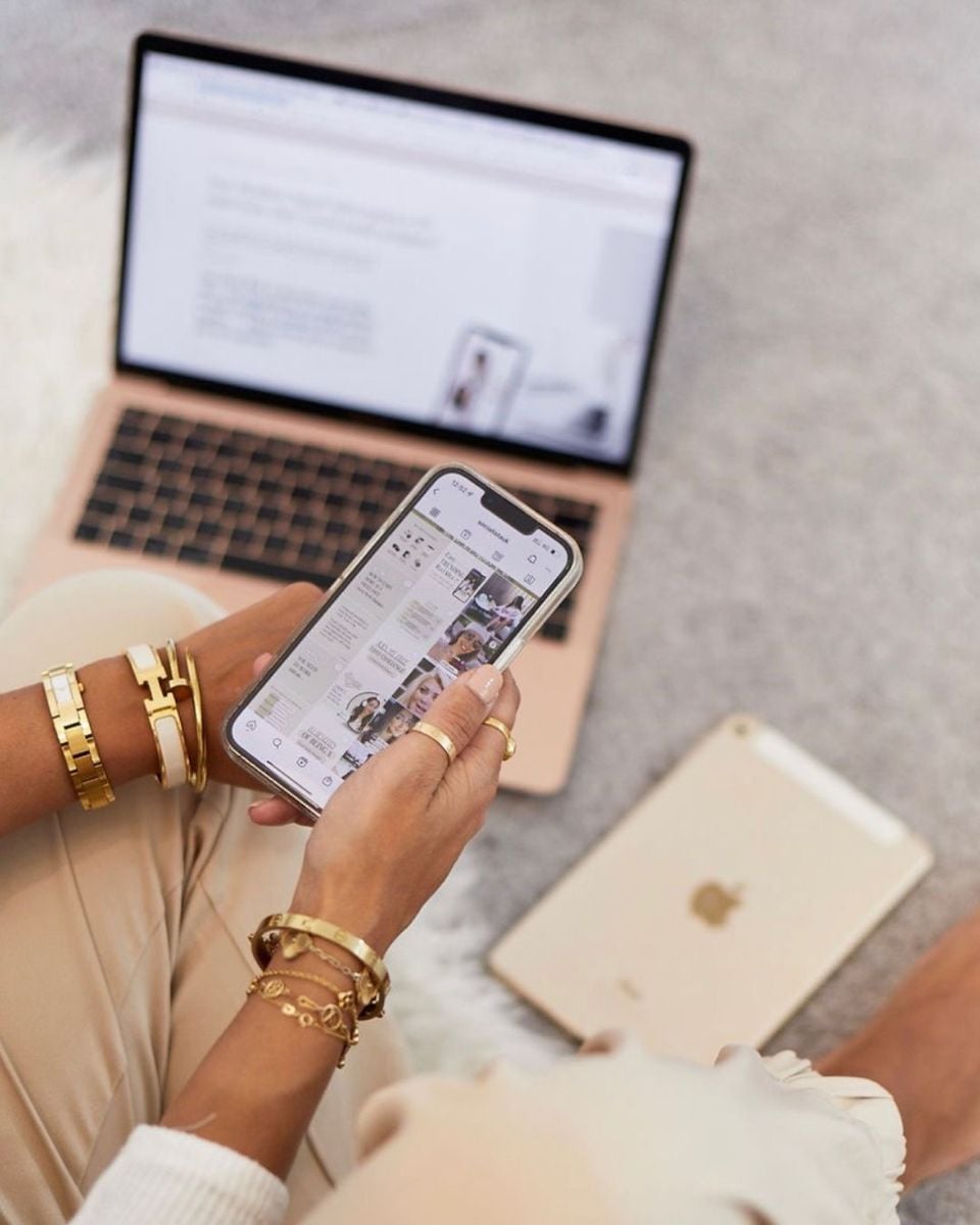 A person wearing gold bracelets holds a smartphone displaying a social media feed. A rose gold laptop and a gold tablet are placed on a soft surface nearby. The setting appears casual and cozy.