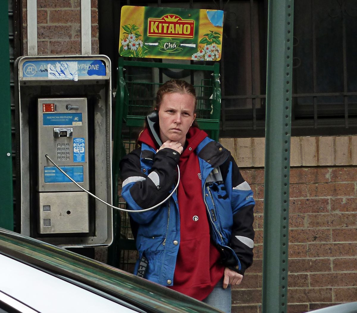 A woman wearing a blue and black jacket over a red hoodie stands at a public payphone, holding the receiver to her ear, with a thoughtful expression. A Kitano advertisement is visible above the phone.