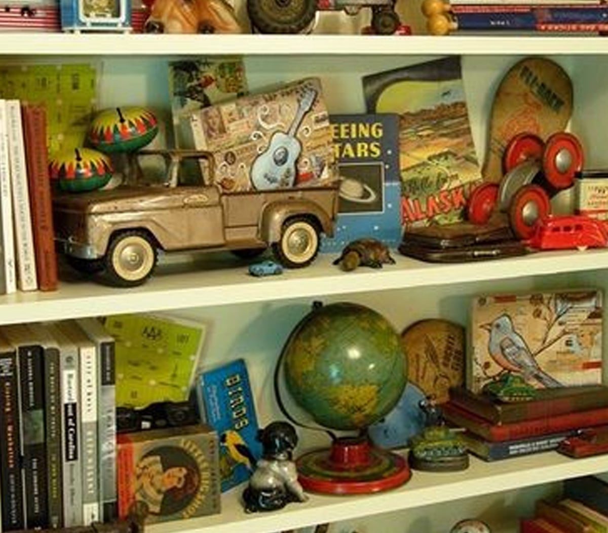 A bookshelf filled with books, a toy truck, maracas, vintage postcards, a globe, toy cars, a duck figurine, and various colorful objects and memorabilia arranged on three shelves.