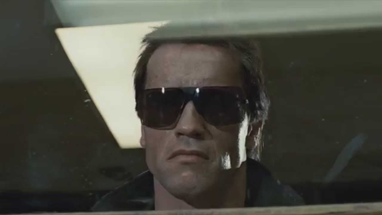 A man with short hair and sunglasses stares forward with a serious expression, viewed through a window with reflections and indoor lighting above him.