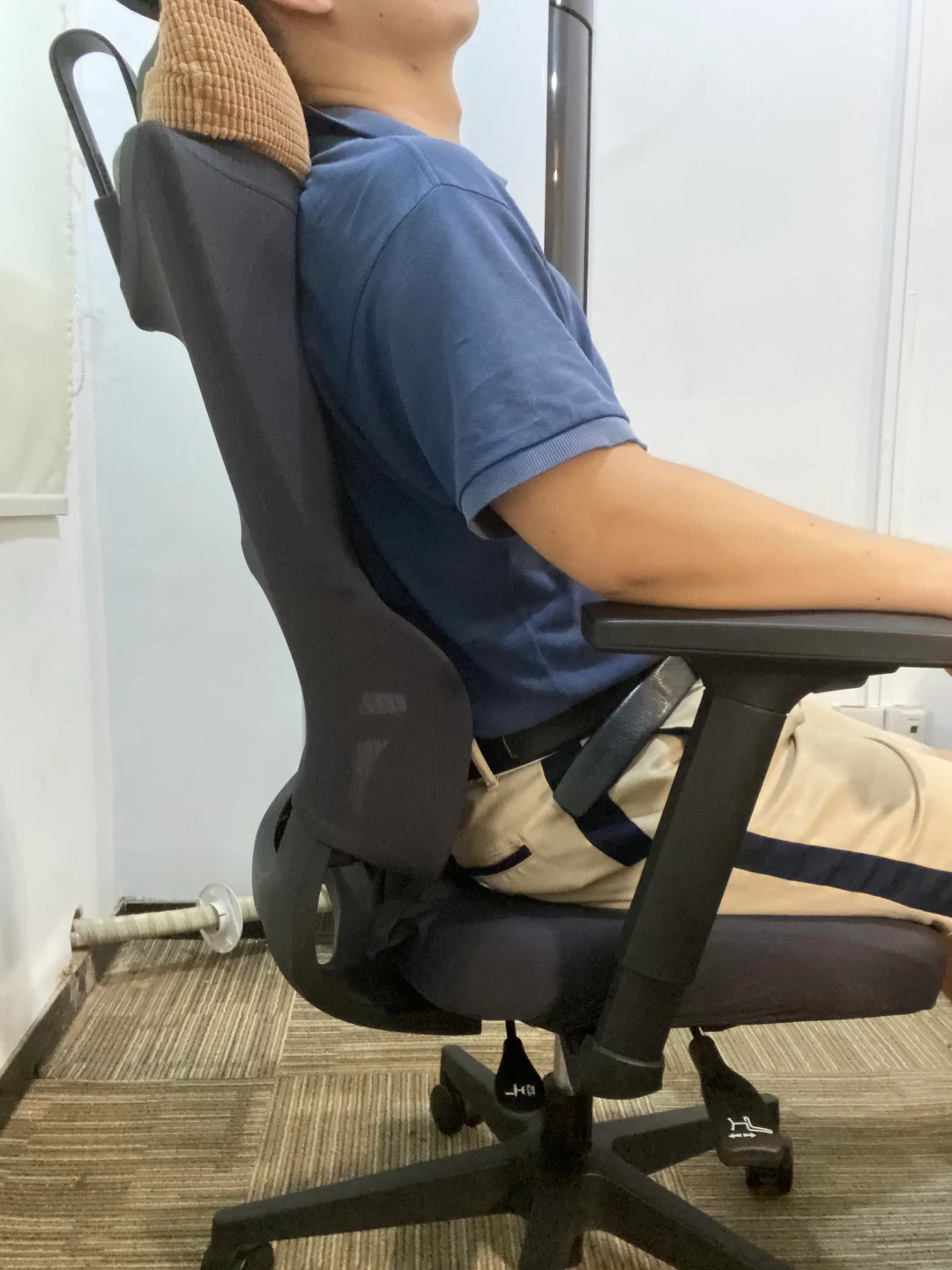 A person wearing a blue shirt and beige pants sits upright in an ergonomic office chair, with a cushion behind their neck. The setting appears to be an office with carpeted flooring.