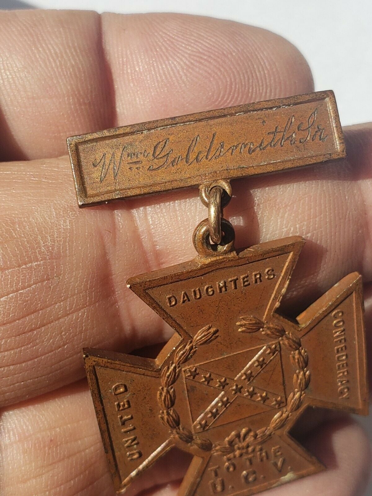 A hand holding a bronze United Daughters of the Confederacy medal with an engraved nameplate reading "Wm. Goldsmith Sr." above a cross-shaped pendant with laurel wreath and text.